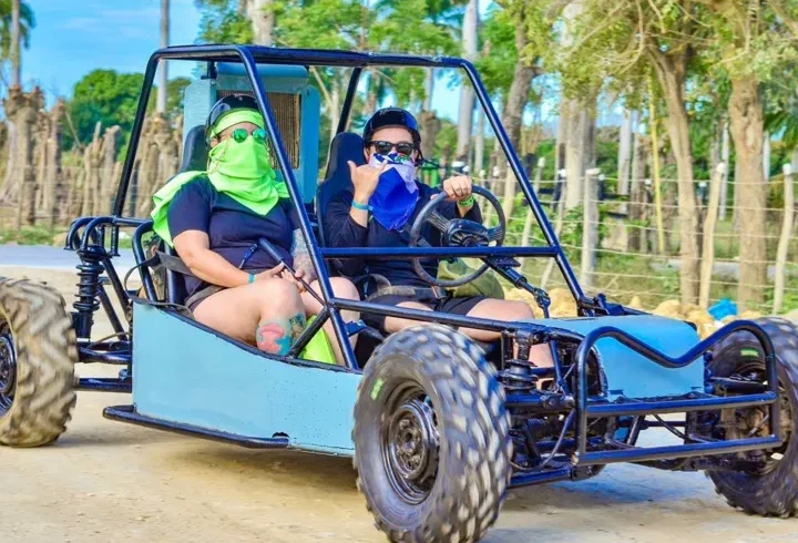 Two people ride in a blue dune buggy on a dirt road during one of the exciting Punta Cana excursions. Both wear sunglasses and bandanas, with the driver making a shaka hand sign. Trees and wooden posts line the background under a bright, blue sky.