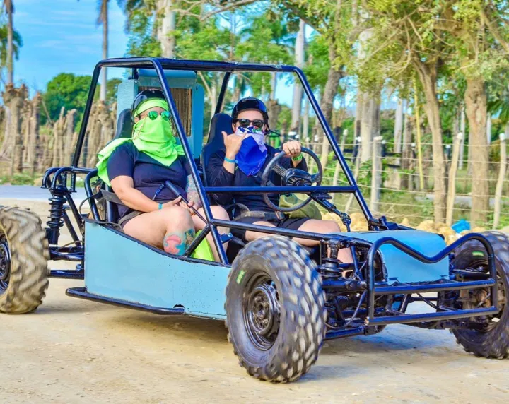Two people ride in a blue dune buggy on a dirt road during one of the exciting Punta Cana excursions. Both wear sunglasses and bandanas, with the driver making a shaka hand sign. Trees and wooden posts line the background under a bright, blue sky.