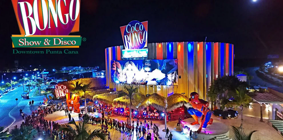 Nighttime aerial view of Coco Bongo Show & Disco in Punta Cana, a must-see on many Punta Cana excursions. The colorful, circular building glows with neon lights as crowds line up outside, palm trees frame the entrance, and a vibrant Coco Bongo sign tops the scene.