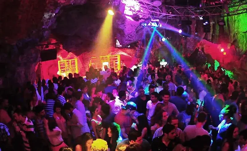 A large crowd dances in a vibrant, rocky cave nightclub filled with colorful lights—purple, blue, green, and yellow. This lively hotspot is a must-see on popular Punta Cana tours for those craving energy and excitement.