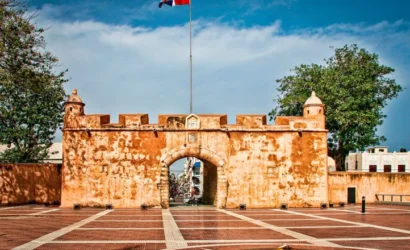 Historic stone archway of the Fortaleza Ozama in Santo Domingo, Dominican Republic, featuring the Dominican flag, surrounded by green trees and a cobblestone plaza, representing cultural heritage and tourism in the Zona Colonial.