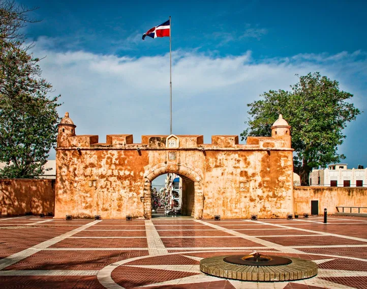 A historic stone fort gate with warm, orange walls stands under a clear blue sky. A Dominican Republic flag flies above—just one of the sights you can explore during unforgettable tours in Punta Cana. The plaza features a monument and flame, flanked by trees.