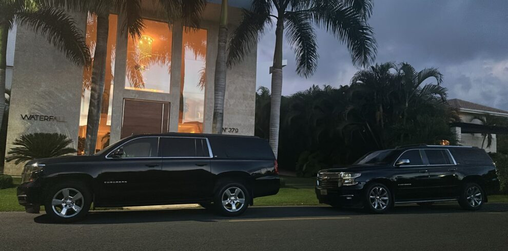 Two large black SUVs are parked side by side at dusk in front of a modern house with tall windows and lush greenery&mdash;reminiscent of exclusive tours in Punta Cana. The warmly lit "WATERFALL" sign enhances the tropical, inviting vibe.