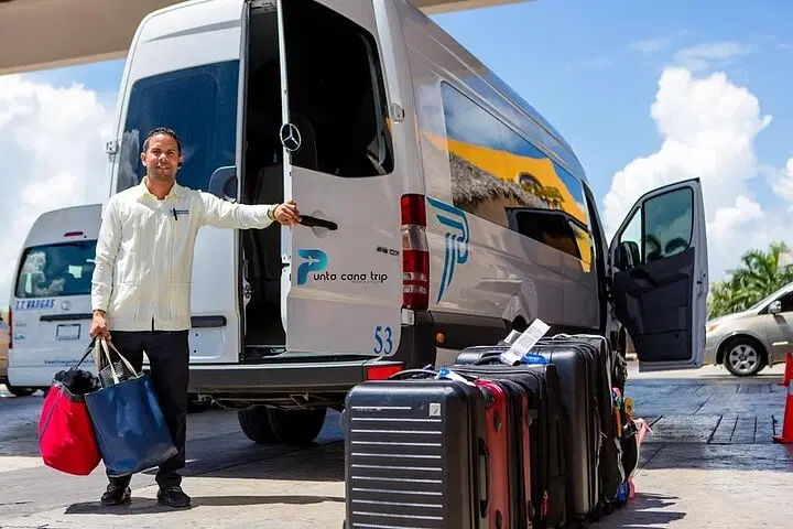A smiling man stands beside an open van door labeled &ldquo;Punta Cana Trip,&rdquo; holding two bags. Suitcases line the pavement, ready for exciting tours in Punta Cana under the sunny blue sky, with another vehicle in the background.