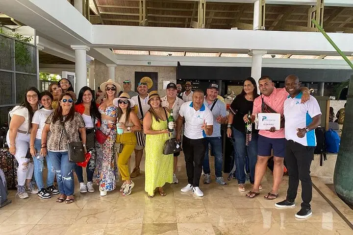 A group of about 20 adults pose indoors at a resort lobby with marble floors, smiling at the camera. Most wear casual summer clothes and sunglasses, some holding drinks&mdash;a fun start to one of their Punta Cana excursions. One man holds a &ldquo;P Quintero&rdquo; sign.