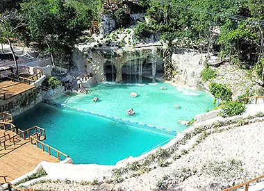 A natural turquoise pool surrounded by rocky cliffs and lush greenery, with small waterfalls flowing in. Wooden walkways border the pool, where a few people float peacefully—an idyllic spot often featured on Punta Cana excursions.