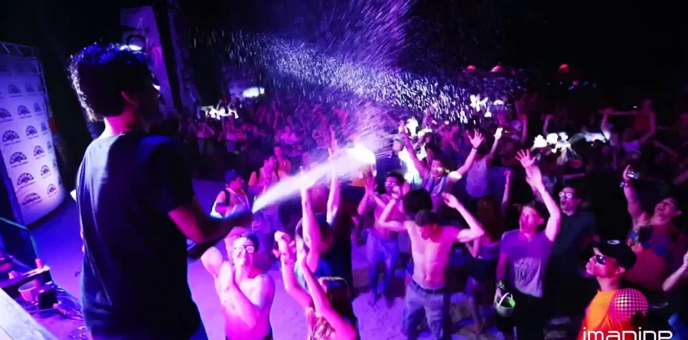 A crowd at a lively outdoor concert in Punta Cana dances and cheers as a performer sprays water over the audience. Colorful lights illuminate the scene, and many shirtless attendees revel in the energetic festival atmosphere.