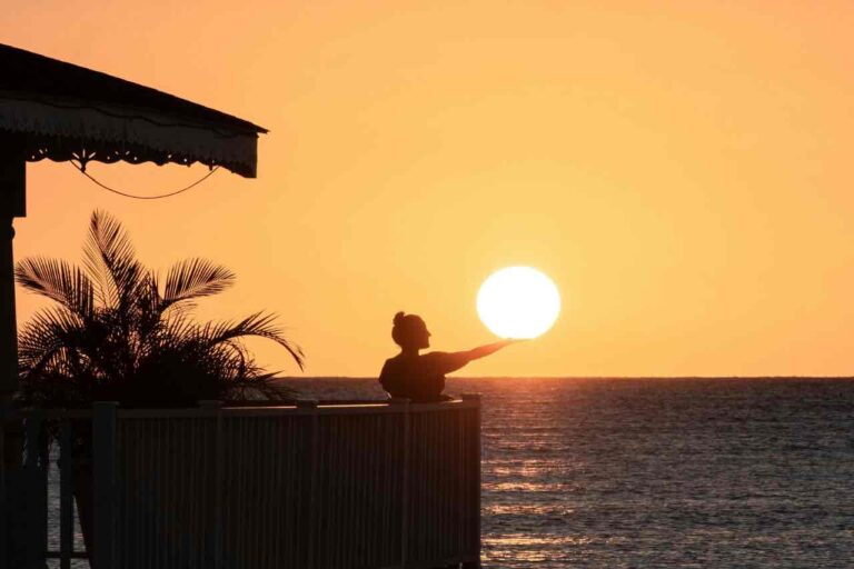 A person stands on a balcony by the sea at sunset, appearing to hold the sun in their outstretched hand. Palm leaves and part of a building are visible on the left, with the sky and water bathed in warm orange light.