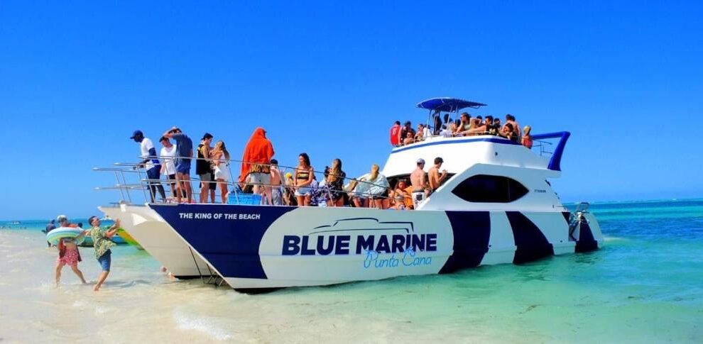 A crowded white and blue catamaran named &ldquo;Blue Marine Punta Cana&rdquo; is anchored in shallow, clear turquoise water. Dozens of people in swimsuits are on the boat and on the beach, enjoying sunny, blue-sky weather. Some walk along the sand; others relax onboard.