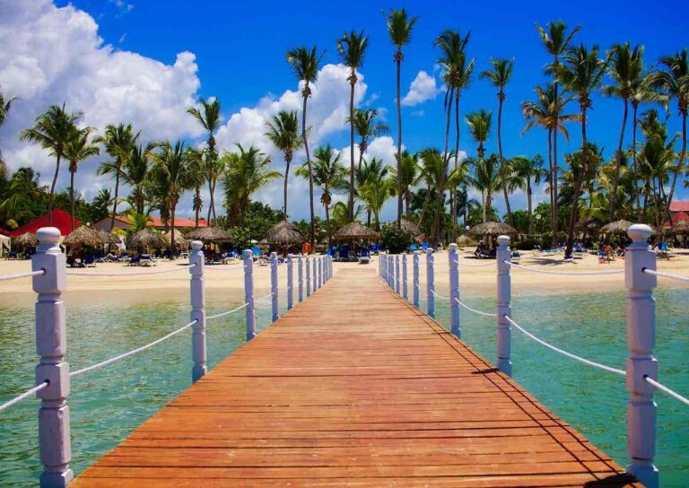A wooden pier extends over clear turquoise water toward a tropical beach lined with tall palm trees and thatched umbrellas. People relax on lounge chairs under the sunny blue sky with scattered white clouds. The scene exudes a vibrant, tropical paradise vibe.