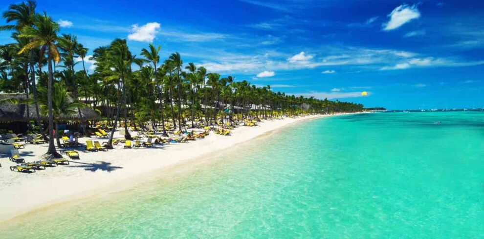 A tropical beach with clear turquoise water, white sand, and rows of palm trees. Yellow lounge chairs and umbrellas line the shore, with people relaxing. The sky is bright blue with a few scattered clouds, creating a vibrant, sunny scene.