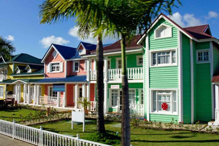 A row of colorful, two-story wooden houses with porches and white railings stands beside a white picket fence. Palm trees and flowers line the sidewalk. The houses are painted green, pink, and blue under a bright, sunny sky with scattered clouds.