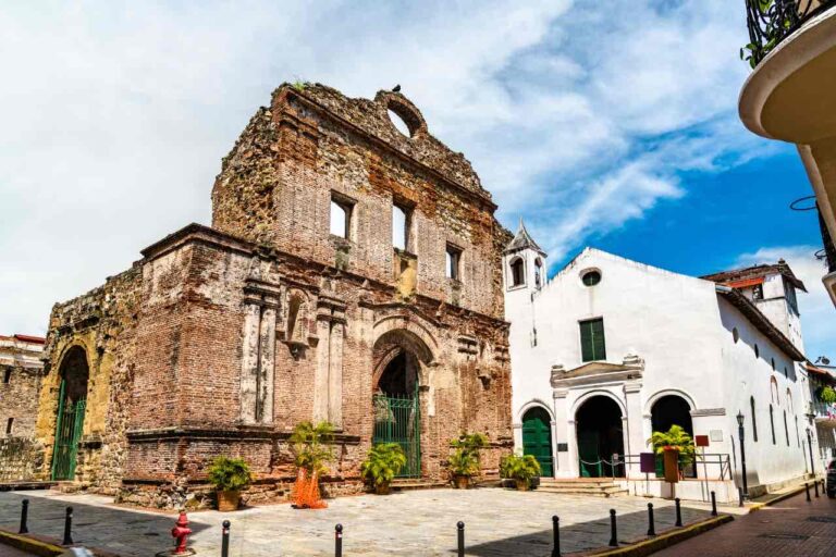 A historic brick church ruin with arched doorways and windows stands next to a white building with a green door and small bell tower, under a partly cloudy blue sky. Potted plants line the stone plaza in front of the structures.
