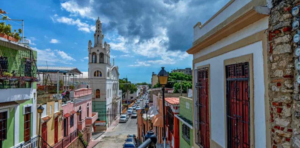 Colorful colonial buildings line a narrow street leading toward a white church with a tower. Cars are parked along the road. A wrought-iron balcony and classic street lamp feature in the foreground. Blue sky with dramatic clouds overhead.