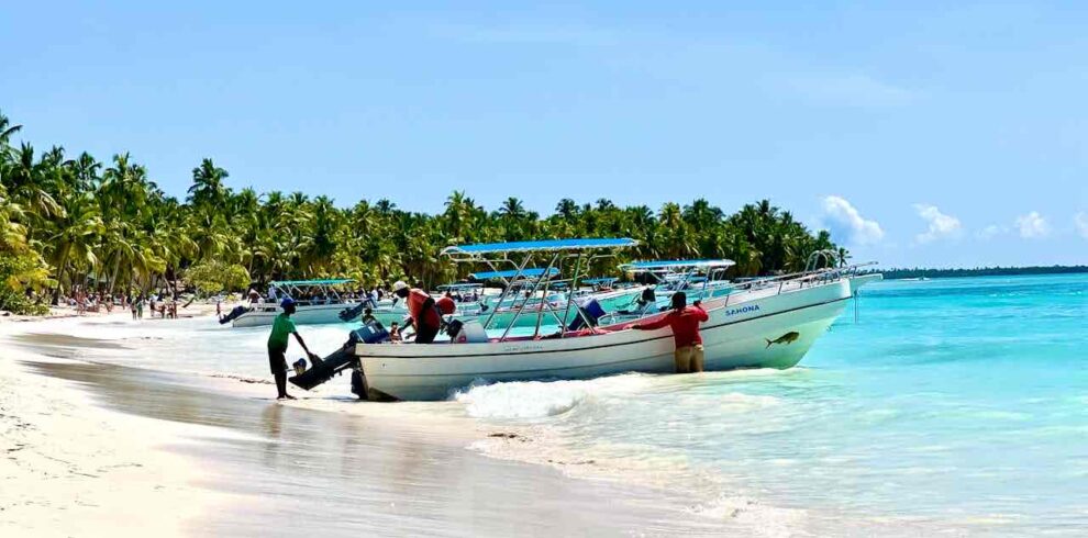 Two motorboats with people onboard rest in shallow turquoise water near a tropical sandy beach lined with palm trees. A man stands next to one boat, touching its motor, while the sky is mostly clear with a few clouds.